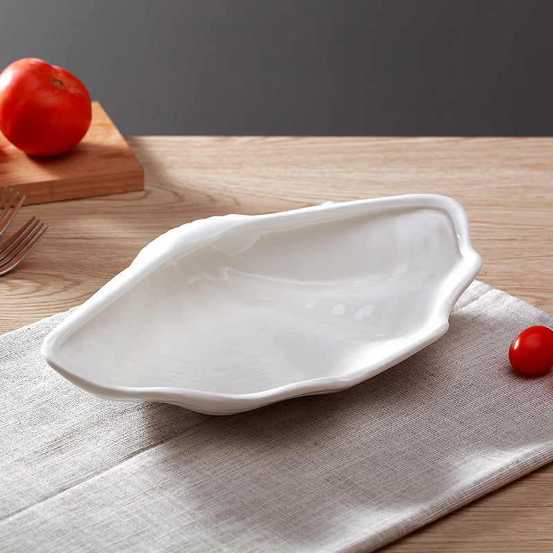 White ceramic shell-shaped plate with irregular scalloped edge, angled view on linen cloth near a tomato