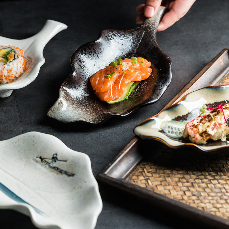 Hand lifting black glazed leaf-shaped ceramic plate topped with salmon, other plates visible nearby