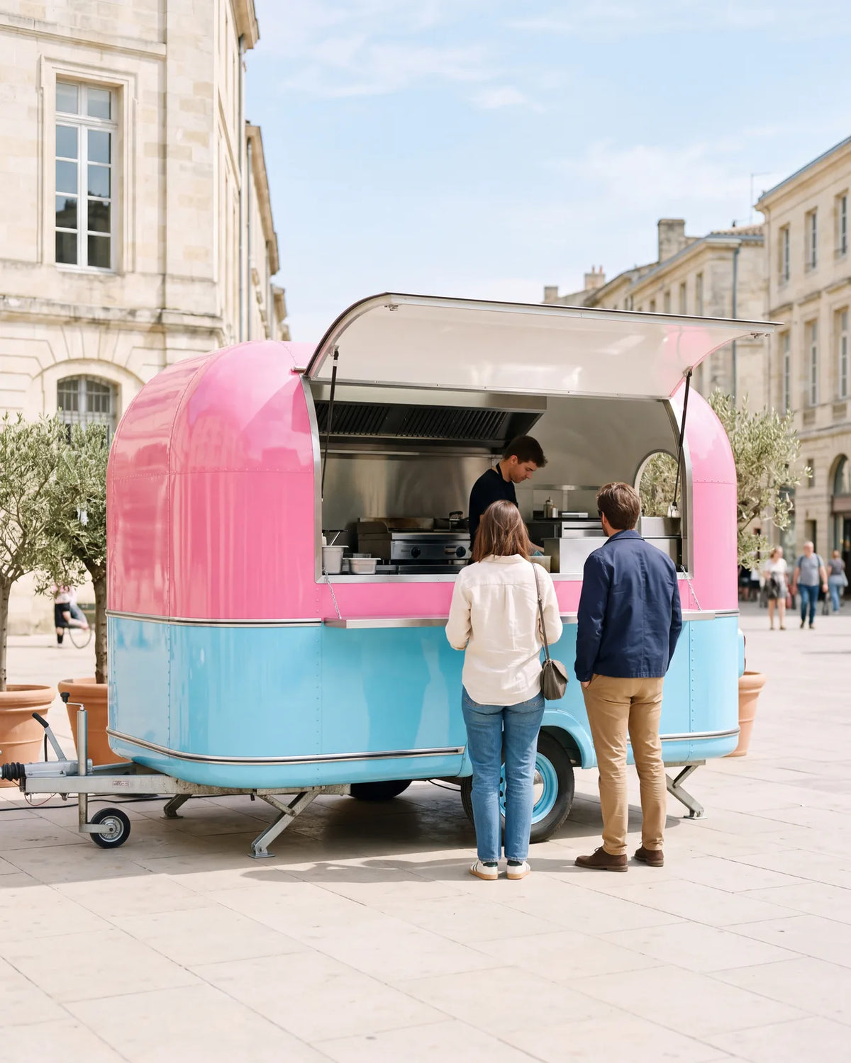 Pink and sky-blue food trailer with its serving hatch open, two customers at the counter on a sunny square