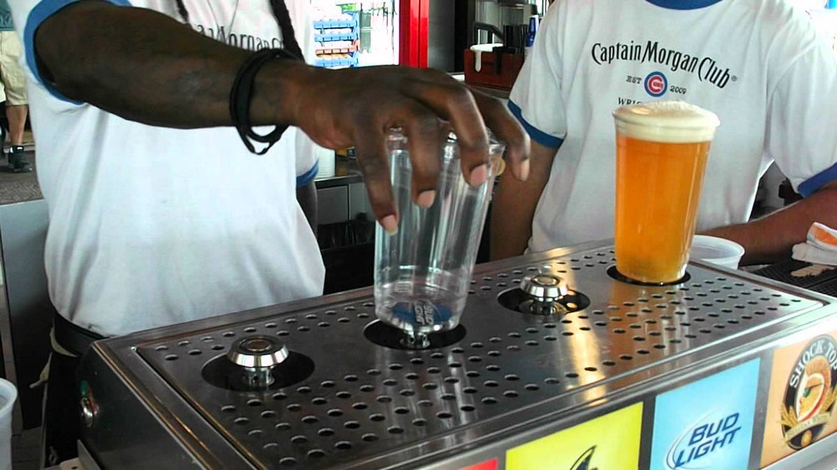 Bar staff lowering two glasses onto the magnetic pads of a bottoms-up dispenser, a poured pint standing beside them