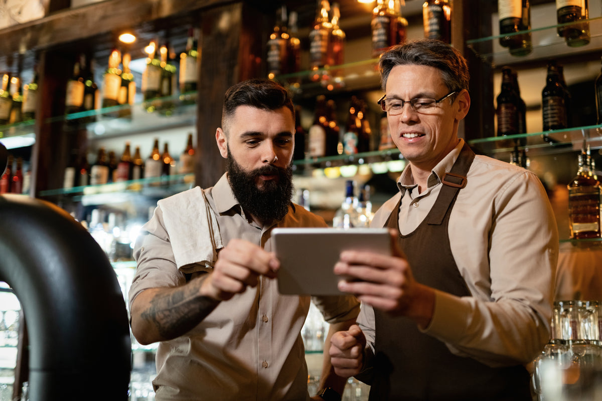 Two bartenders looking at a tablet together behind a lit back bar of bottles