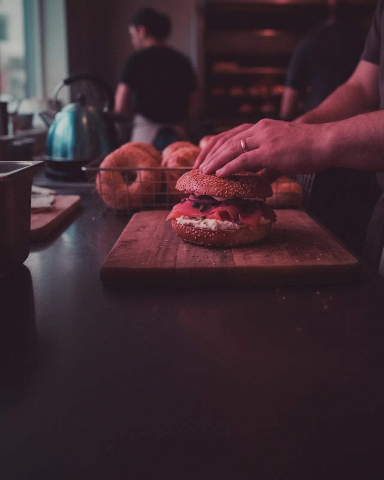 The filled bagel seen close on its board across the counter, the bagel basket and the shop kitchen soft behind
