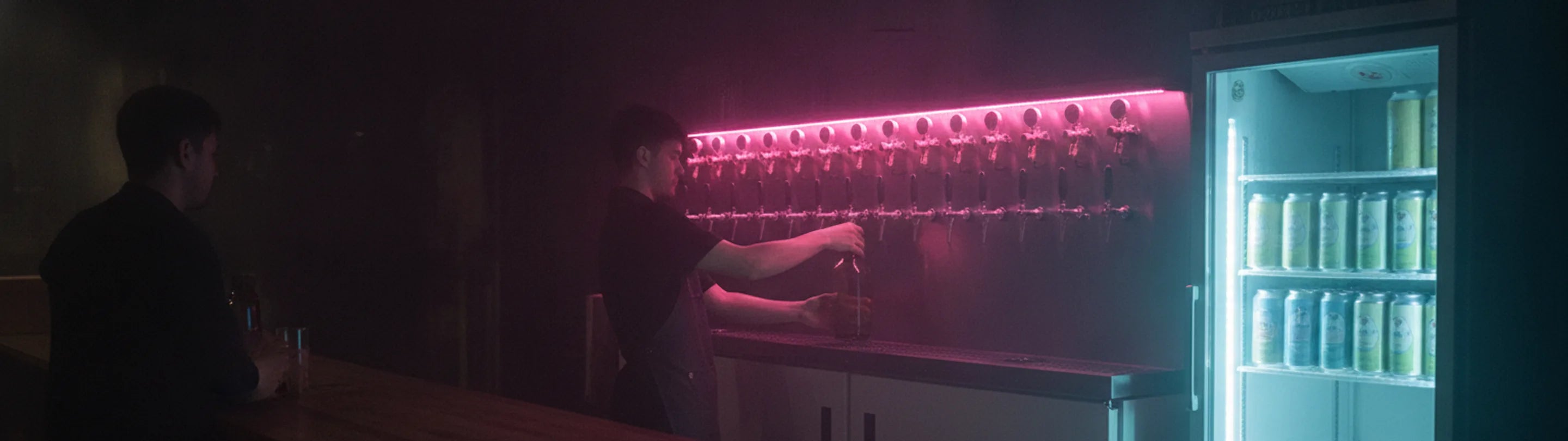 Beer refill shop, a staff member filling a bottle from a twelve-tap wall, a glass-door can fridge beside it and a customer at the counter
