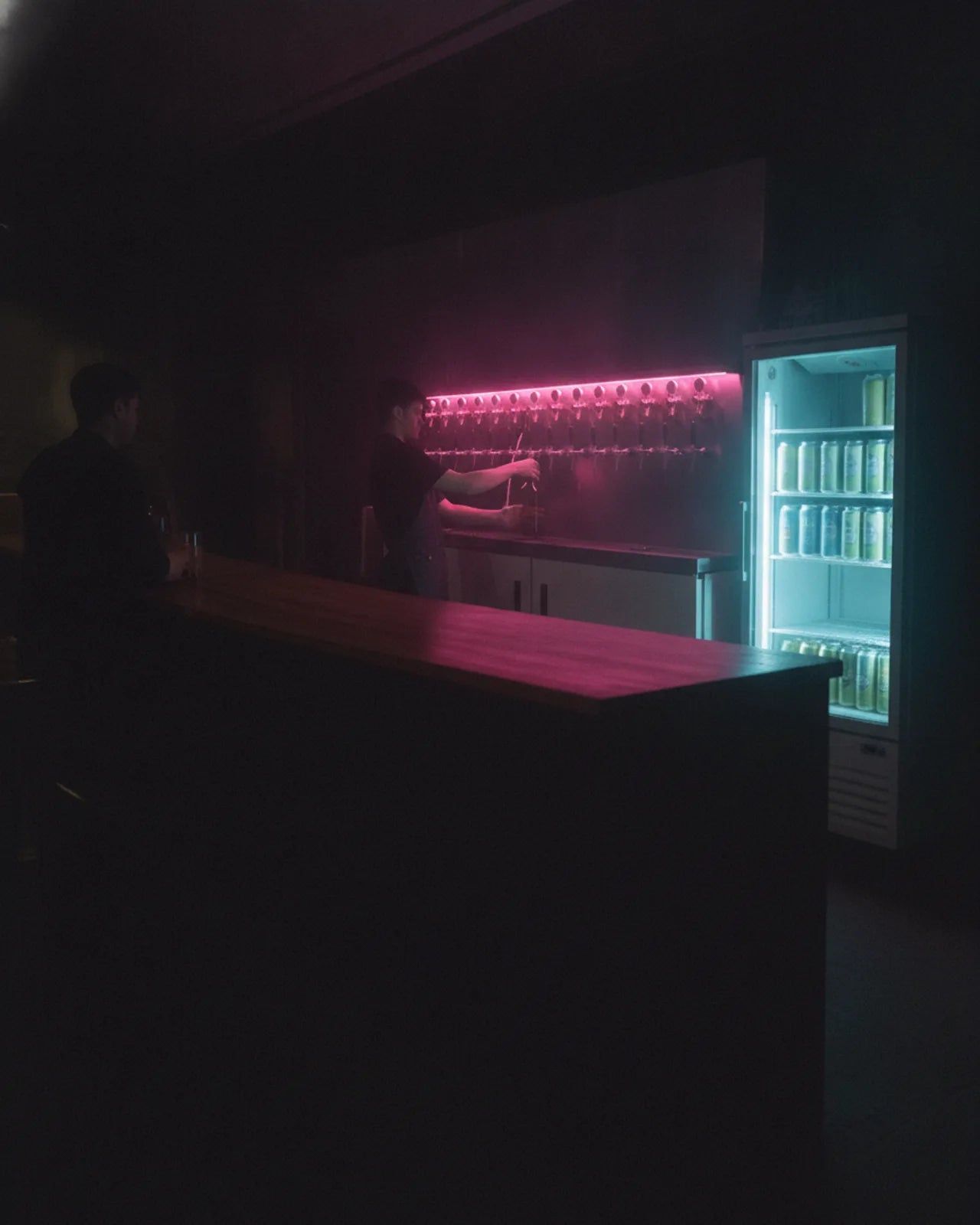 Beer refill bar seen from the counter, a lit twelve-tap wall and a glass-door fridge of cans