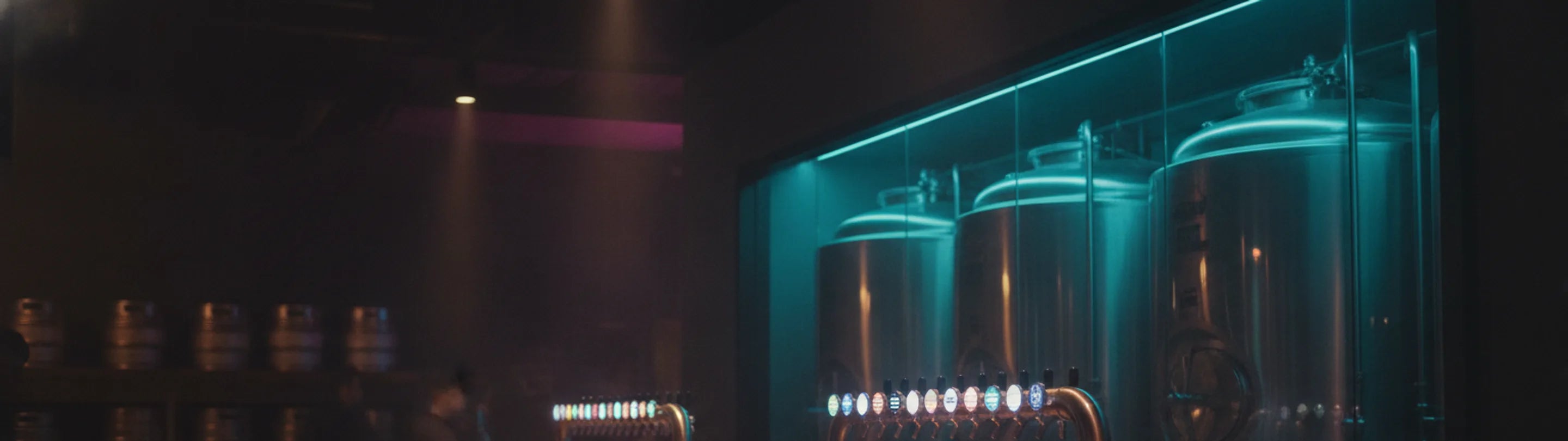 Stainless conditioning tanks lit cyan behind a glass partition, a row of taps on the bar in front, stacked kegs receding into darkness on the left