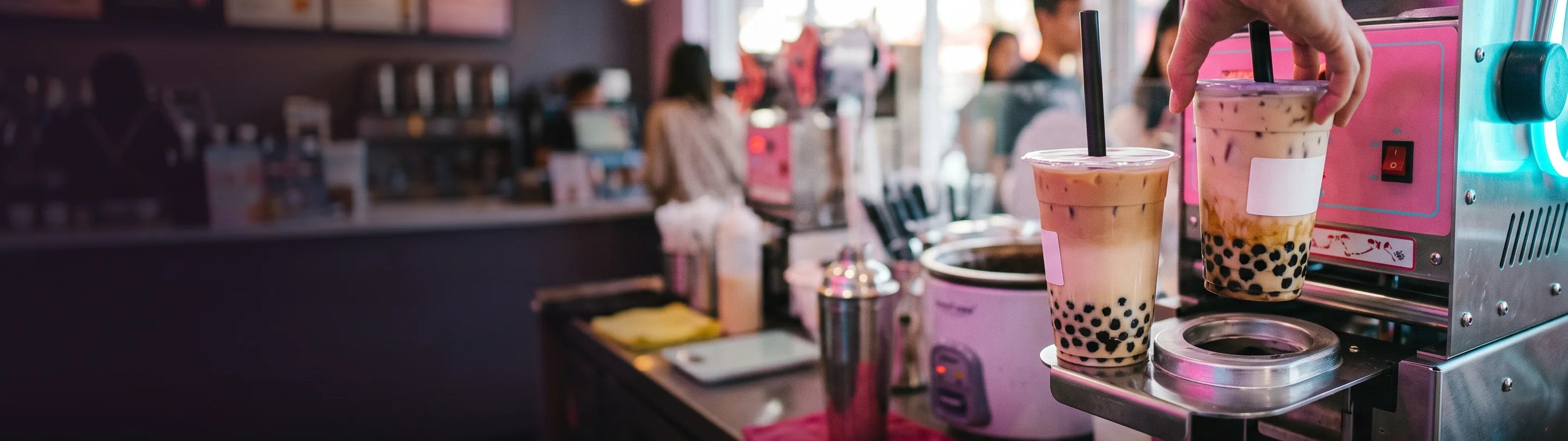 Two clear cups of milk tea with tapioca pearls settled in the base on the tray of a stainless cup-sealing machine, a hand lifting one, the shop counter behind