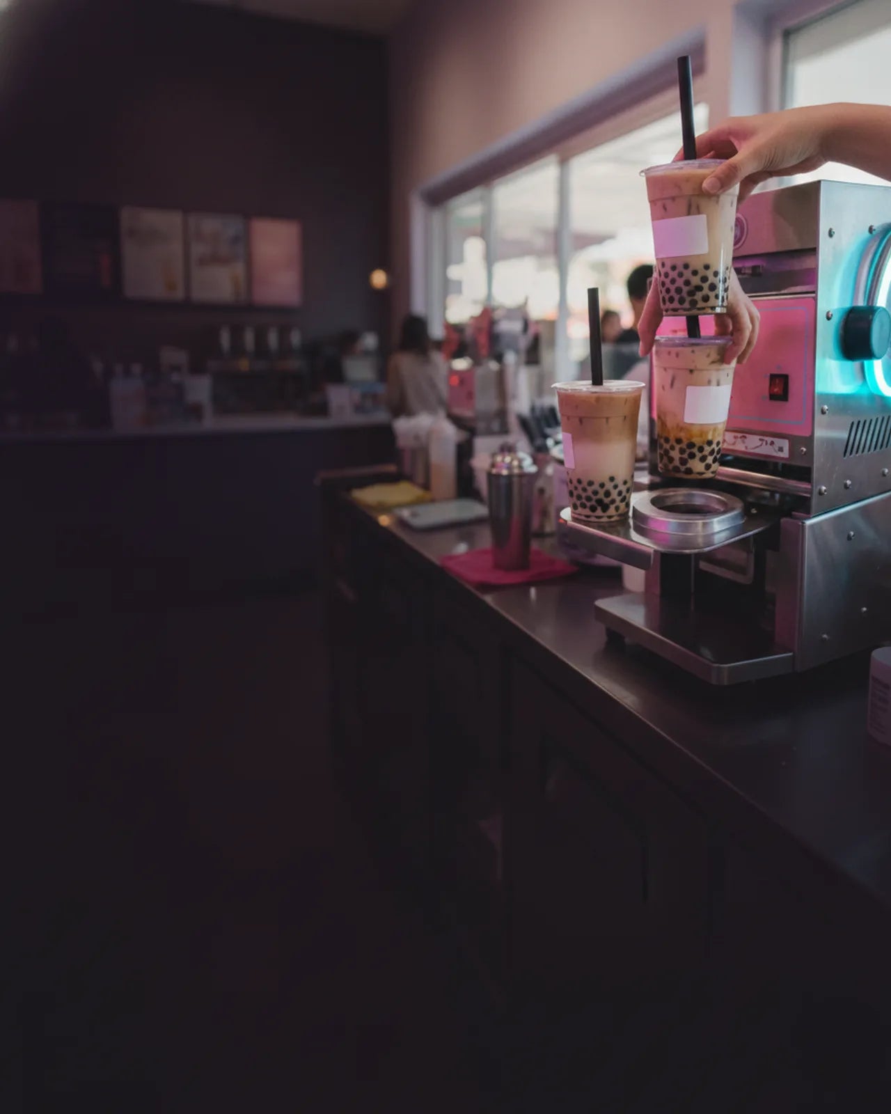 Three cups of milk tea on the sealing machine tray seen from the guest side of the counter, the menu boards and open shopfront out of focus behind