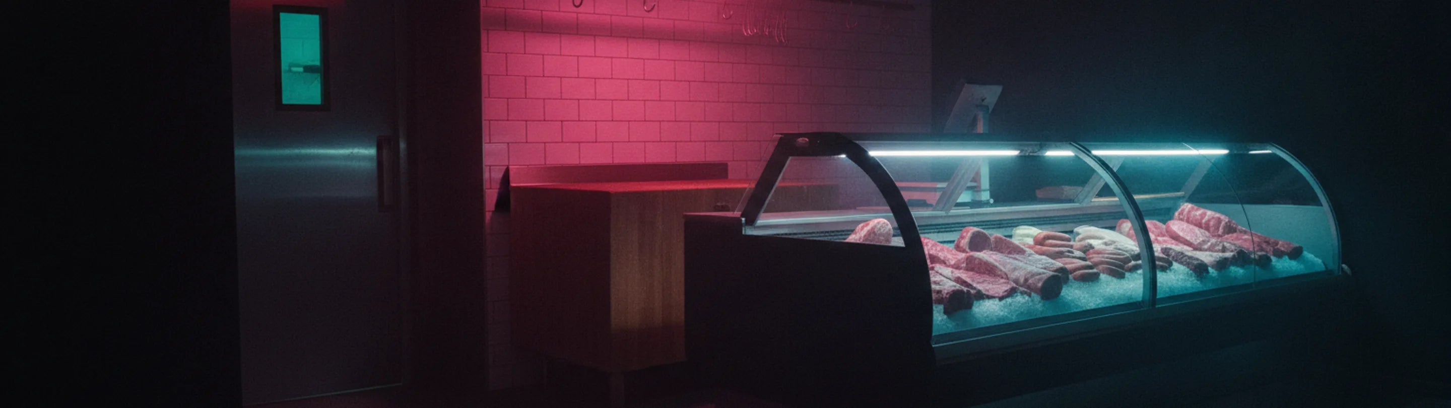 A curved-glass refrigerated counter of cuts laid on ice under cold light, tiled wall and cold-room door receding into shadow on the left
