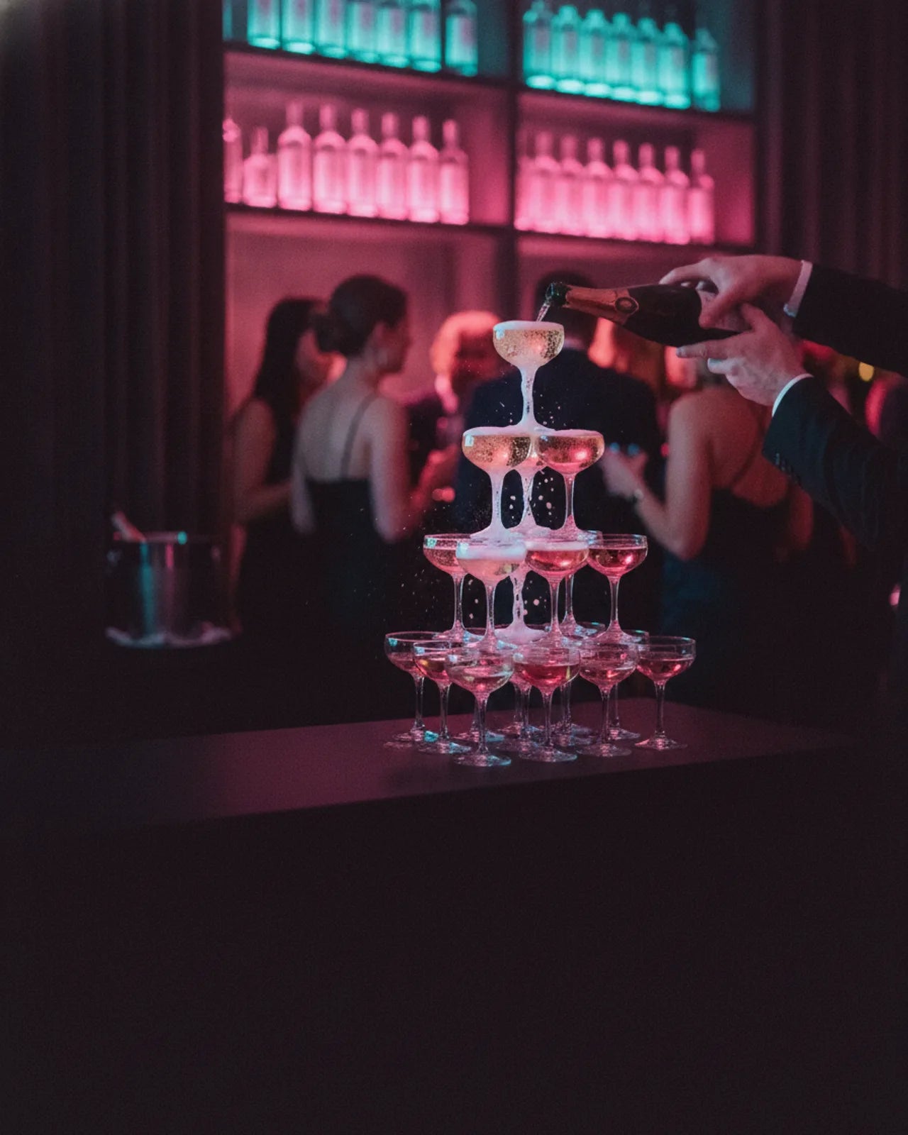 Champagne poured into the top glass of a coupe tower on a bar counter, lit bottle shelves behind