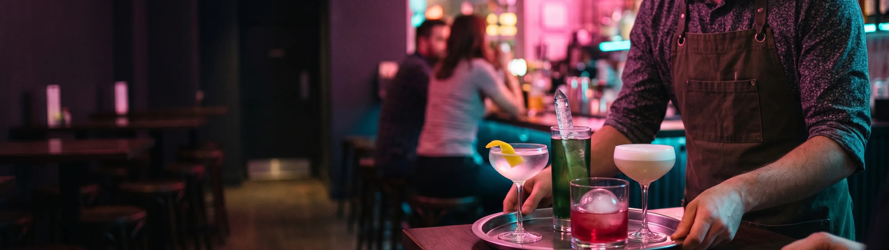Bartender carrying a tray of four cocktails from the bar, seated guests and back bar blurred behind