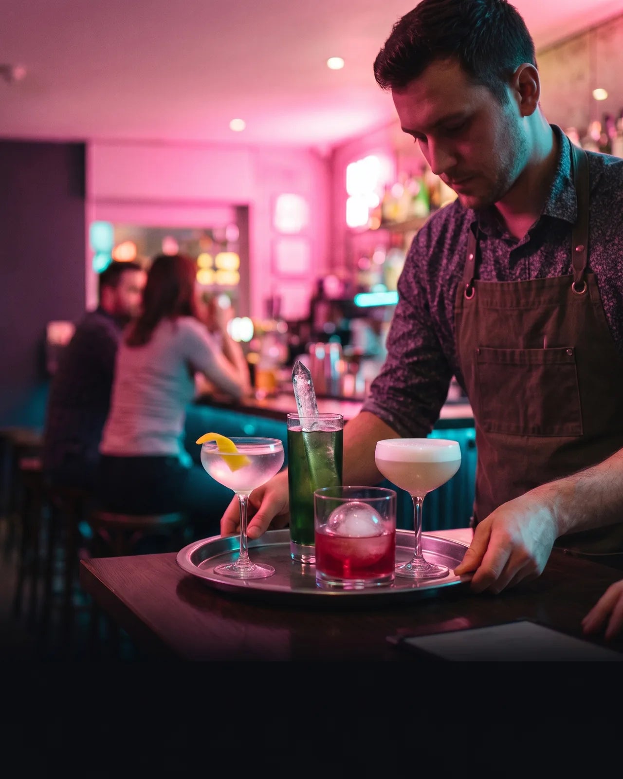 A bartender in an apron setting down a tray of four cocktails — a coupe with a lemon twist, a tall green highball with a clear ice spear, a rocks glass with an ice sphere and a foamed sour — on the end of the bar, guests seated along it behind