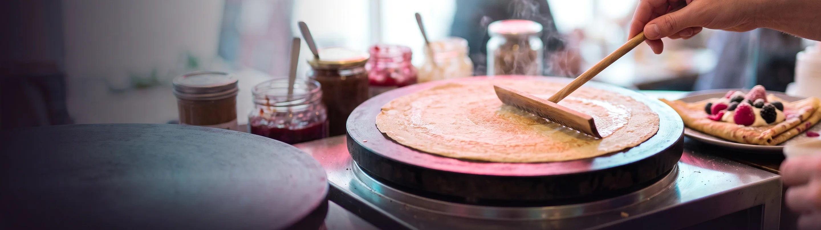 A crepe cooking on a round cast-iron griddle plate being spread thin with a wooden rateau, glass jars of jam and chocolate spread lined up behind, a second griddle in the foreground
