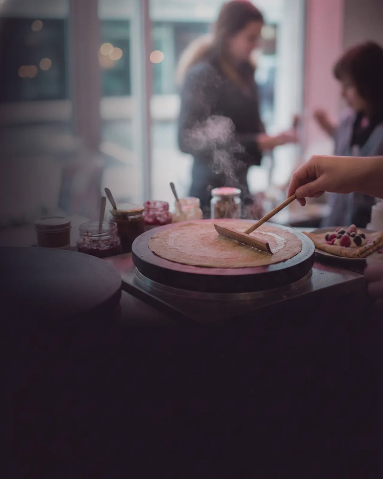 The griddle plate close from the guest side, steam rising off the crepe as the rateau passes, filling jars and a finished plate behind