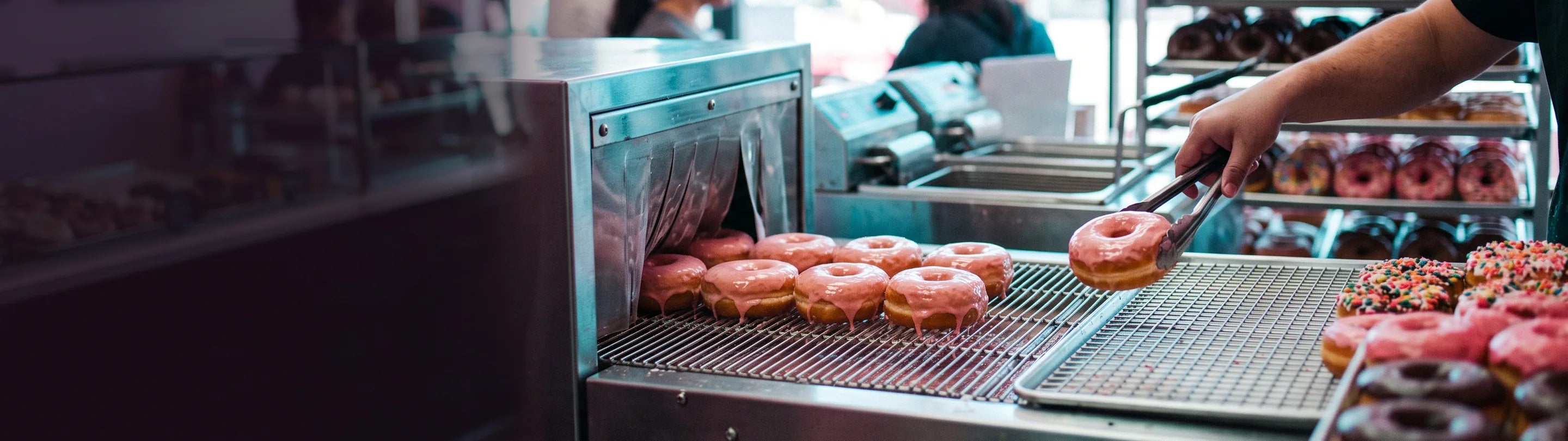 Pink-glazed ring donuts coming out of a stainless glazing machine onto a wire draining rack, a hand lifting one with tongs, trays of sprinkled and chocolate donuts on the right