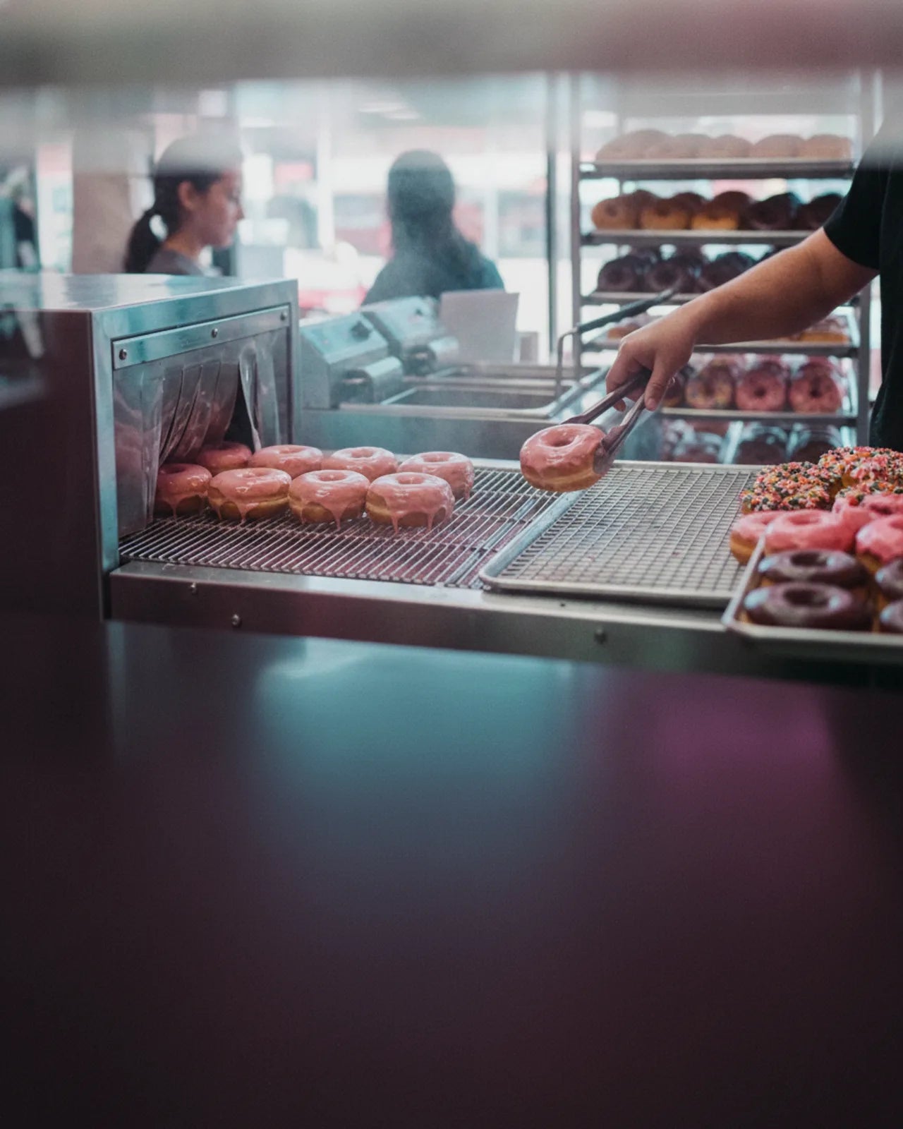 The glazing machine mouth close from the counter side, a row of glazed donuts on the rack and racks of finished trays behind the staff