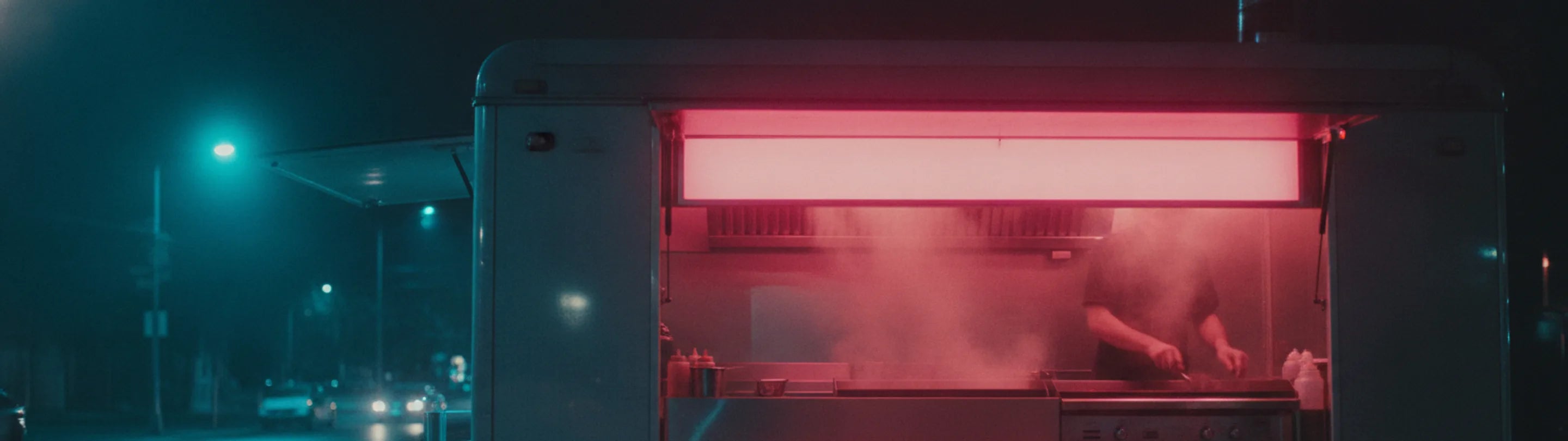 A food truck serving hatch open and lit from inside at night, steam rising from the griddle, wet street reflections receding on the left