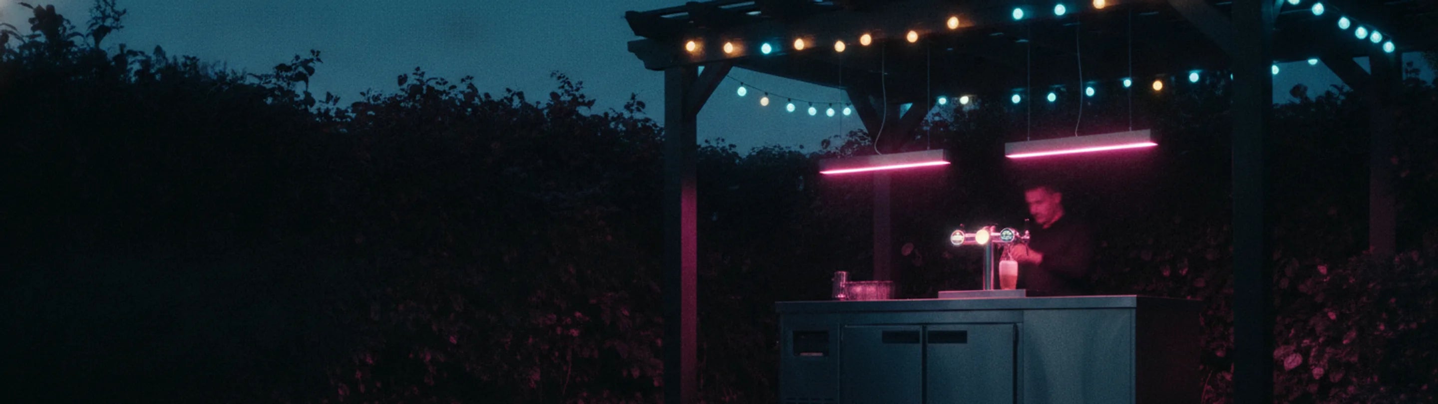 A garden bar at dusk: a stainless refrigerated bar cart under a timber pergola, a two-tap beer cooler pouring into a frosted glass, pink linear pendants and a string of warm bulbs overhead, dark hedge behind