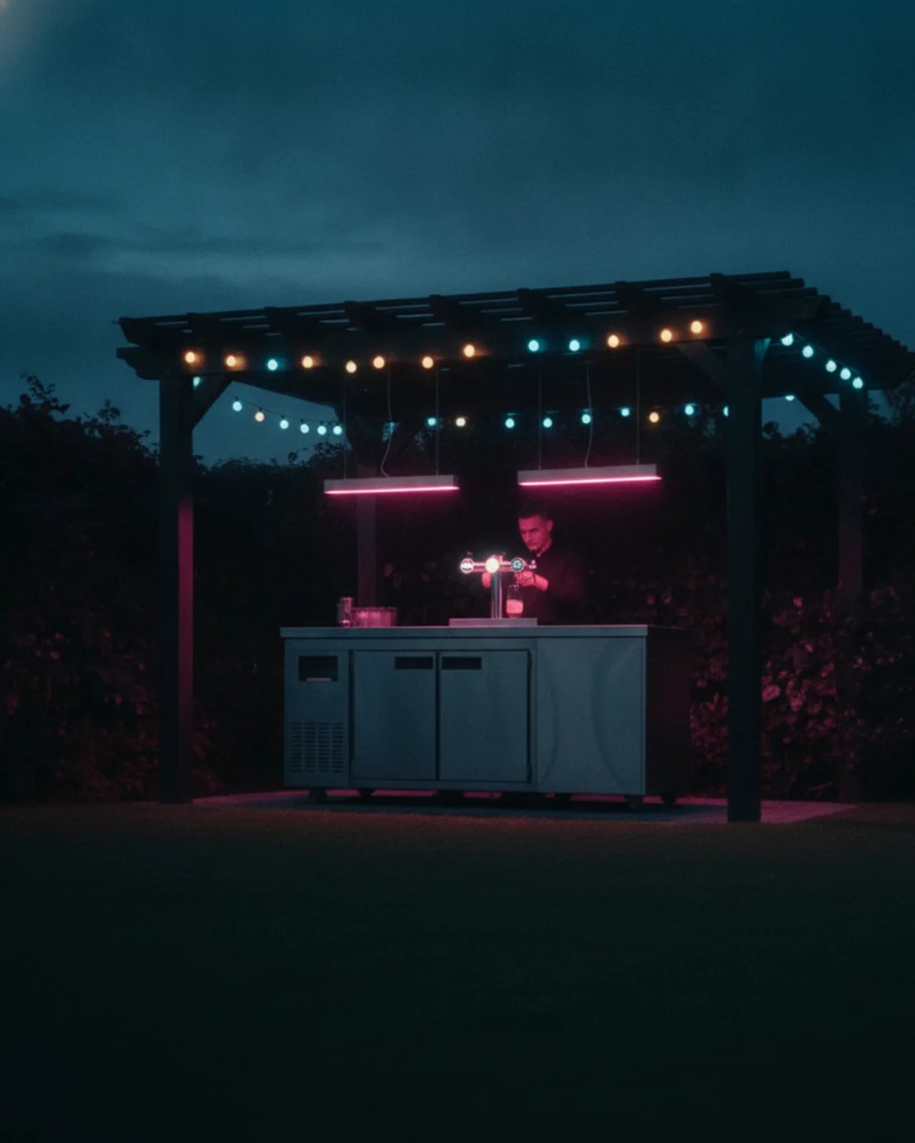 Vertical view of the garden bar: the two-tap cooler on the stainless cart, pendant light above and the lit pergola beam over a dark lawn