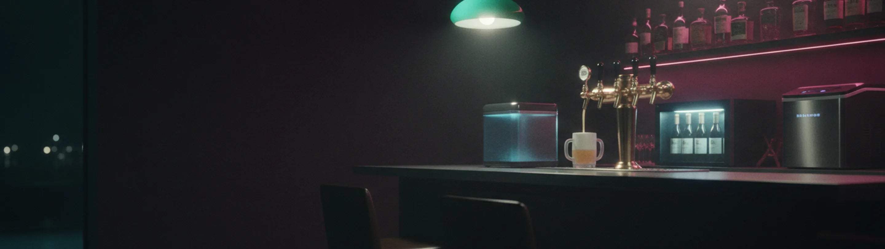 A home bar at night: a four-tap gold draft tower pouring into a frosted glass, a glass froster and a lit four-bottle wine dispenser along the counter, backlit whiskey shelf behind, a green pendant lamp overhead