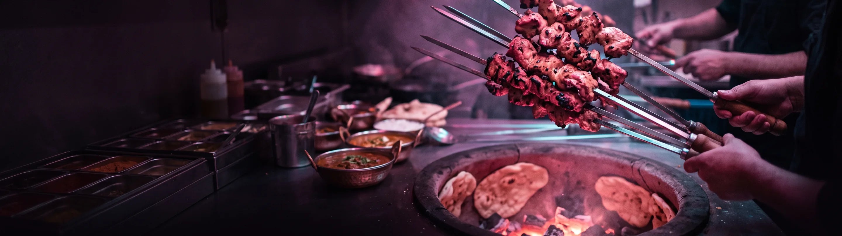 Chef reaching into a lit tandoor oven with skewers, naan on the rim, restaurant kitchen behind