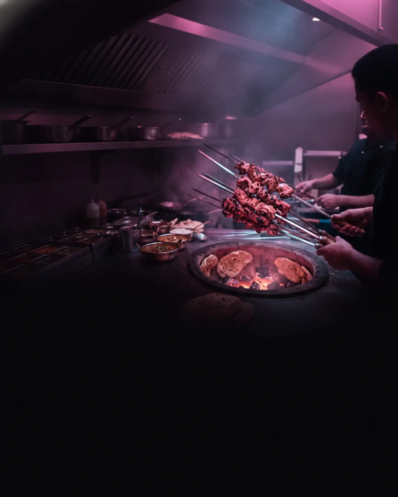 Chef lifting loaded skewers from a lit clay tandoor, naan breads baking on the oven rim