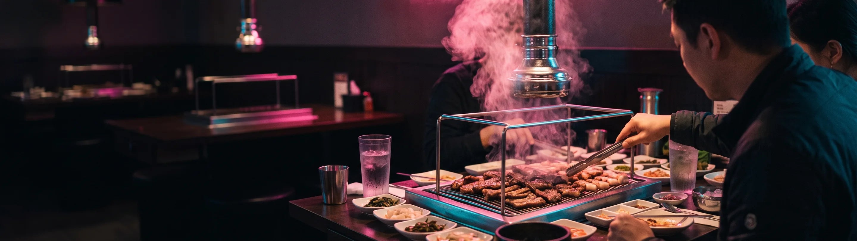 A table grill loaded with pork belly under a polished stainless extraction duct pulling the smoke up, small white banchan dishes crowding the table edge, two diners in profile