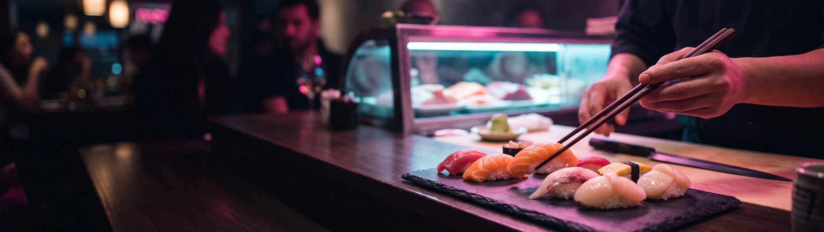Chef slicing fish behind a refrigerated sushi counter, nigiri plated on the pass