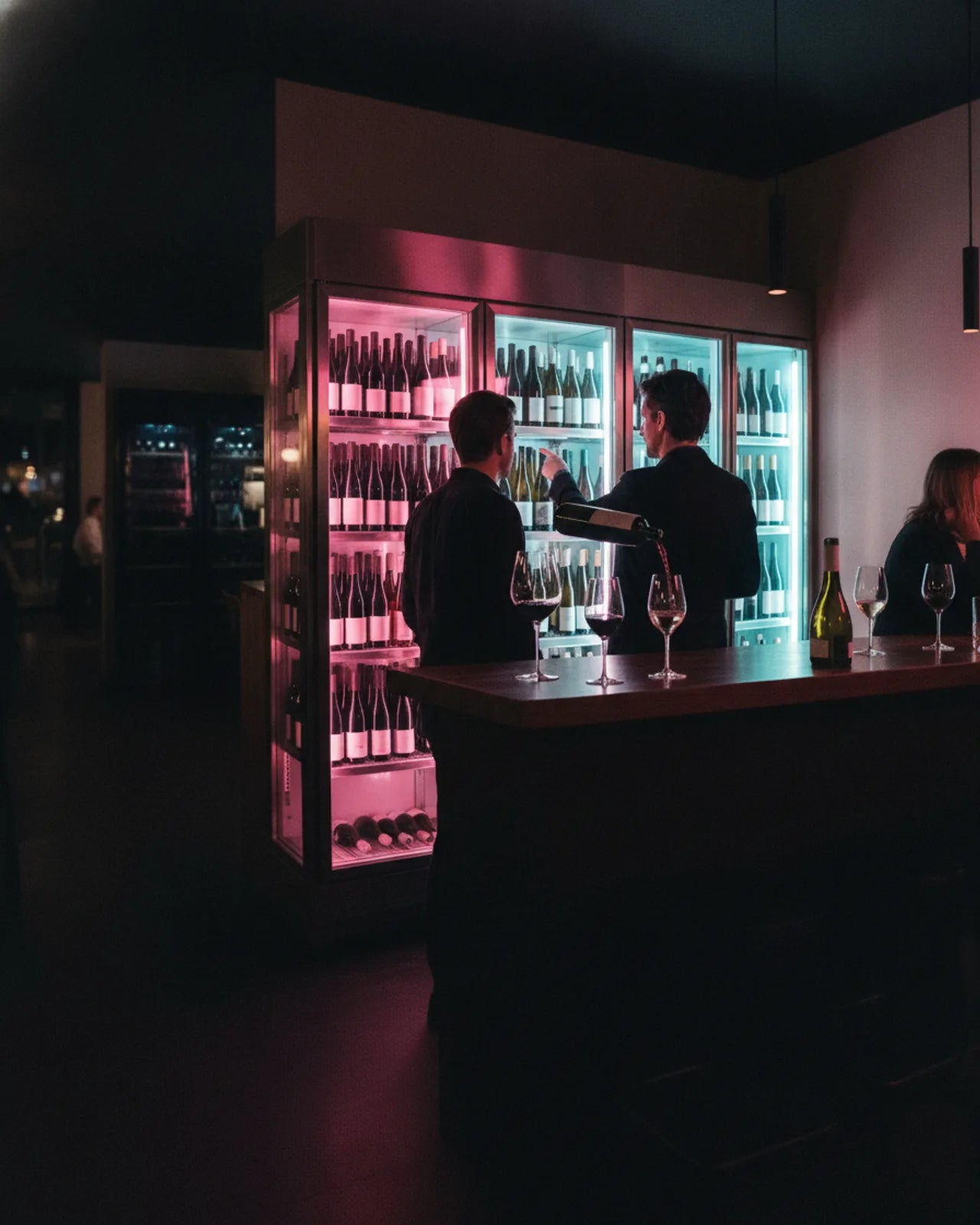 Bartender pouring red wine at a wine bar counter, backlit fridges of bottles glowing behind