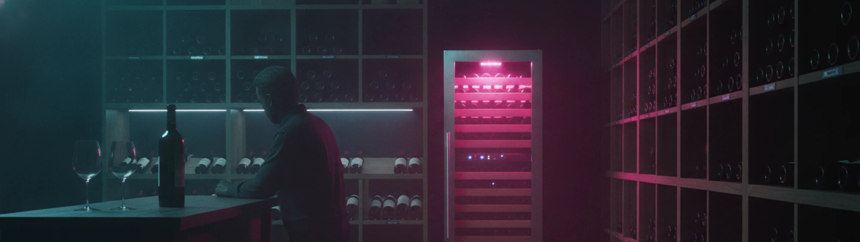 Wine shop at night, a dual-zone glass-door wine refrigerator glowing pink between wooden bottle racks, a man at a tasting counter with two glasses and a bottle