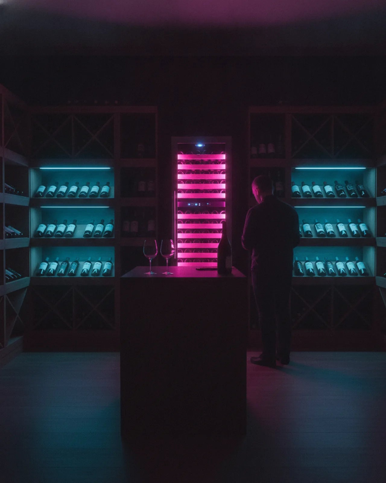 Wine shop tasting counter with two glasses in front of a lit dual-zone wine refrigerator, bottle shelves lit cyan on both sides